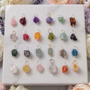 May include: A collection of twenty-four raw gemstone pendants arranged in four rows on a white marble square. Each pendant features a unique, irregularly shaped crystal in various colors including pink, red, purple, green, blue, and orange, attached to a small metal jump ring.