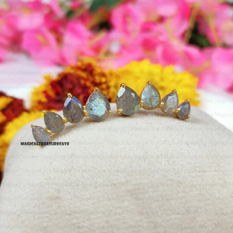 May include: A pair of gold-toned ear climbers featuring teardrop-shaped labradorite stones. The stones are a mix of gray and blue hues, set in a delicate, curved design. The earrings are displayed on a neutral surface with a blurred floral background.