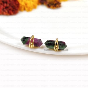 Può includere: Un paio di orecchini con pietre preziose. Ogni orecchino presenta un cristallo esagonale verde e magenta avvolto con filo dorato. Gli orecchini sono presentati su una superficie bianca, con accenti sfocati arancioni e gialli sullo sfondo.