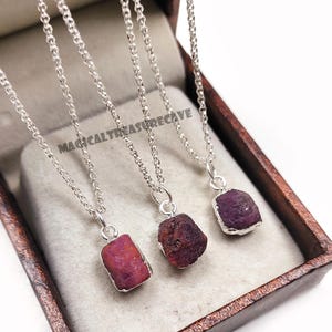 Puede incluir: Tres collares de plata con colgantes de rubí en bruto. Los colgantes son de varios tonos de rojo y morado. Los collares se exhiben en una caja de madera con el texto "MAGICALTREASURECAVE".