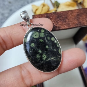 May include: A silver pendant featuring a green and black oval stone. The stone has a speckled pattern with white spots. The pendant is in a small wooden box.
