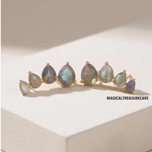 May include: A pair of gold-tone ear climbers featuring a row of four teardrop-shaped labradorite gemstones in each piece, displaying iridescent blue and grey flashes. The text MAGICALTREASURECAVE is printed in black near the right earring.