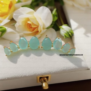 May include: A pair of gold earrings with a curved design featuring five teardrop-shaped light blue gemstones.