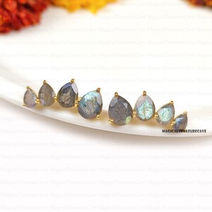 May include: A pair of gold-toned earrings featuring teardrop-shaped labradorite stones. The stones display a mix of gray and iridescent blue-green hues. The earrings are arranged in a curved line, showcasing a unique design.