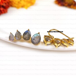 May include: A pair of gold-colored ear climbers featuring four pear-shaped labradorite stones. The stones display a mix of gray, blue, and iridescent colors. The earrings are displayed on a white surface.