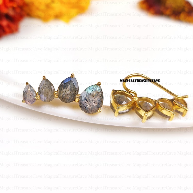 May include: A pair of gold-colored ear climbers featuring four pear-shaped labradorite stones. The stones display a mix of gray, blue, and iridescent colors. The earrings are displayed on a white surface.