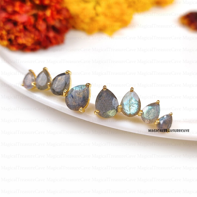 May include: Earrings featuring teardrop-shaped labradorite stones set in gold-colored metal. The stones display a mix of gray, blue, and iridescent hues. The earrings are arranged on a white surface. The text "MAGICALTREASURECAVE" is visible.