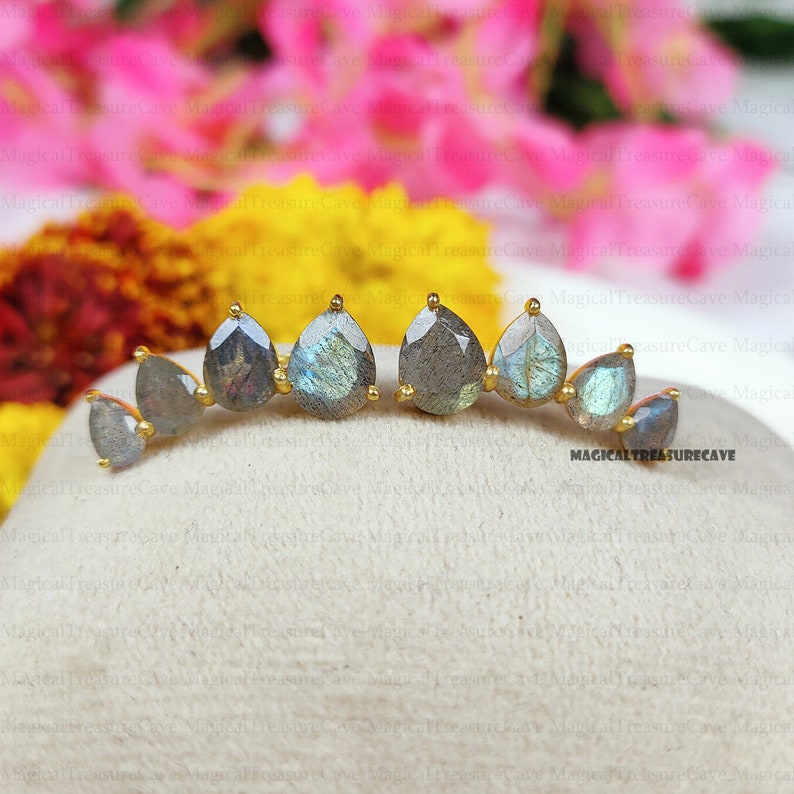 May include: A pair of gold-toned ear climbers featuring teardrop-shaped labradorite stones. The earrings have a curved design with multiple stones, each set in a gold bezel. The labradorite stones display a mix of gray and blue hues.