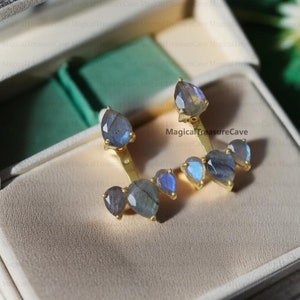Peut inclure: Une paire de boucles d'oreilles dorées avec plusieurs pierres précieuses bleu-gris irisées en forme de goutte. Les boucles d'oreilles ont un design unique, avec les pierres précieuses disposées en grappe. Les boucles d'oreilles sont présentées dans un écrin crème.