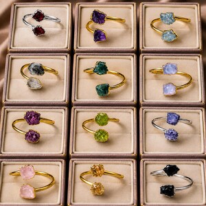 May include: A collection of twelve open-band rings, each featuring two raw, uncut gemstone points in various colors including red, purple, blue, green, pink, yellow, and black, set on either silver-toned or gold-toned metal bands in individual boxes.