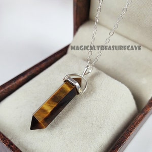 May include: A silver necklace with a pendant made of tiger's eye gemstone. The pendant is a brown and gold hexagonal shape. The necklace is displayed inside a brown jewelry box. The text "MAGICALTREASURECAVE" is visible.
