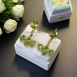Puede incluir: Un par de pendientes de oro con piedras preciosas de peridoto verde. Los pendientes están diseñados con una piedra en forma de lágrima en la parte superior y tres piedras más pequeñas en la parte inferior. Los pendientes se muestran en una caja blanca.