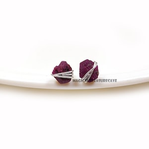 May include: Two raw ruby gemstone earrings. Each ruby is a deep magenta color, wrapped with silver-colored wire. The earrings are displayed on a white, slightly curved surface. The text "MAGIC TREASURECAVE" is visible.
