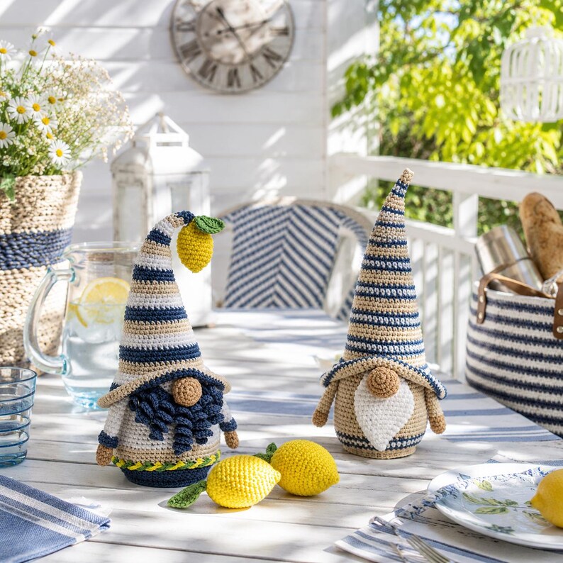 May include: Two crochet gnomes with beige, blue, and white striped hats and beards sit on a table with lemons. One gnome holds a lemon. The scene is set on a sunny porch with a clock and a basket of flowers in the background.