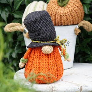 May include: A crocheted pumpkin gnome wearing a black hat with a green and yellow ribbon. The gnome is sitting on a white stool with a bucket behind it.
