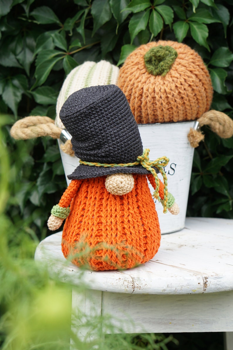 May include: A crocheted pumpkin gnome wearing a black hat with a green and yellow ribbon. The gnome is sitting on a white stool with a bucket behind it.