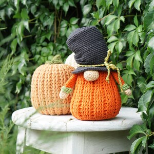 May include: A crocheted gnome wearing a black hat and holding a crocheted orange pumpkin. The gnome and pumpkin are sitting on a white stool.