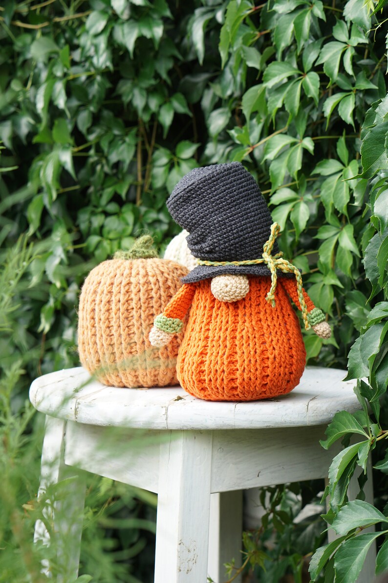 May include: A crocheted gnome wearing a black hat and holding a crocheted orange pumpkin. The gnome and pumpkin are sitting on a white stool.