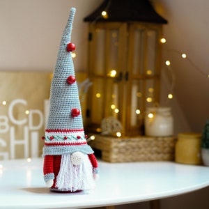 May include: A gray and red crocheted gnome ornament with a white beard and red hat. The gnome is standing on a white table with a wooden background.