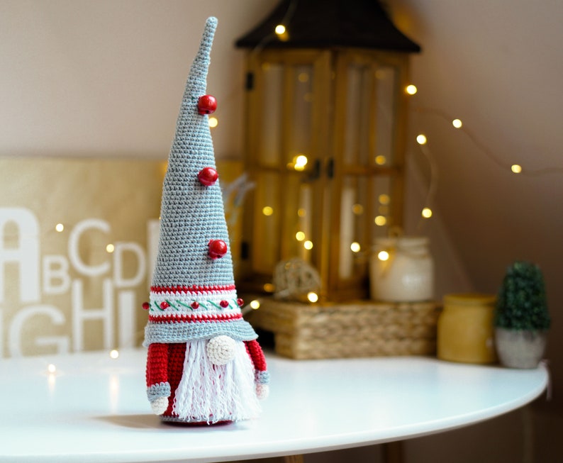 May include: A gray and red crocheted gnome ornament with a white beard and red hat. The gnome is standing on a white table with a wooden background.