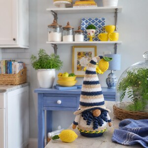 May include: A handmade crochet gnome with a striped hat in navy, beige, and white, holding a lemon. The gnome has a white body, blue beard, and a yellow lemon. The gnome is a decorative item.