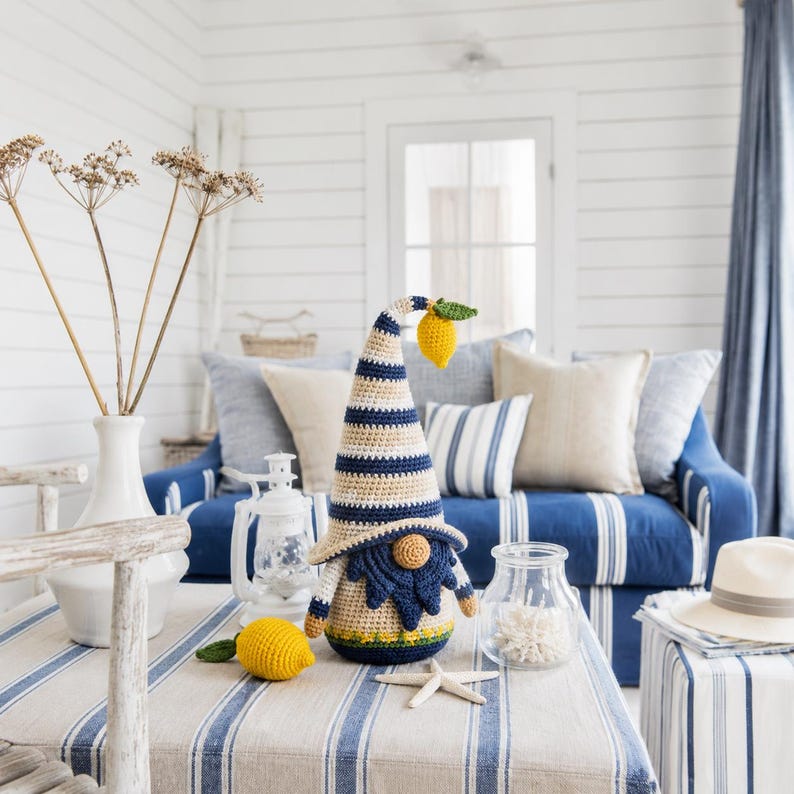 May include: A crocheted gnome with a striped blue and beige hat and a yellow lemon accent. The gnome has a blue beard and is placed on a striped blue and white table. A yellow crocheted lemon is next to the gnome. The background includes a blue sofa and white walls.