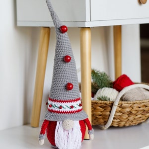 May include: A gray and red crocheted gnome with a white beard and red pom-poms on its hat. The gnome is sitting on a white surface.