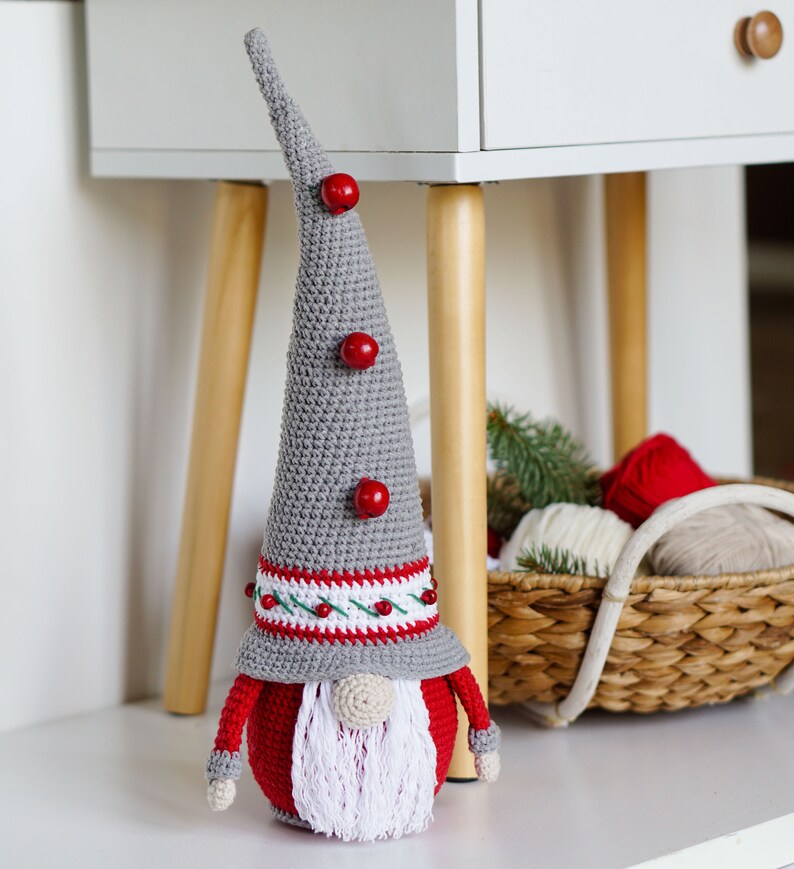 May include: A gray and red crocheted gnome with a white beard and red pom-poms on its hat. The gnome is sitting on a white surface.