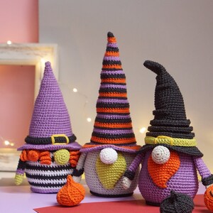 May include: Three crocheted Halloween gnomes with purple, orange, and black hats. The gnomes are holding pumpkins and are standing on a red surface.