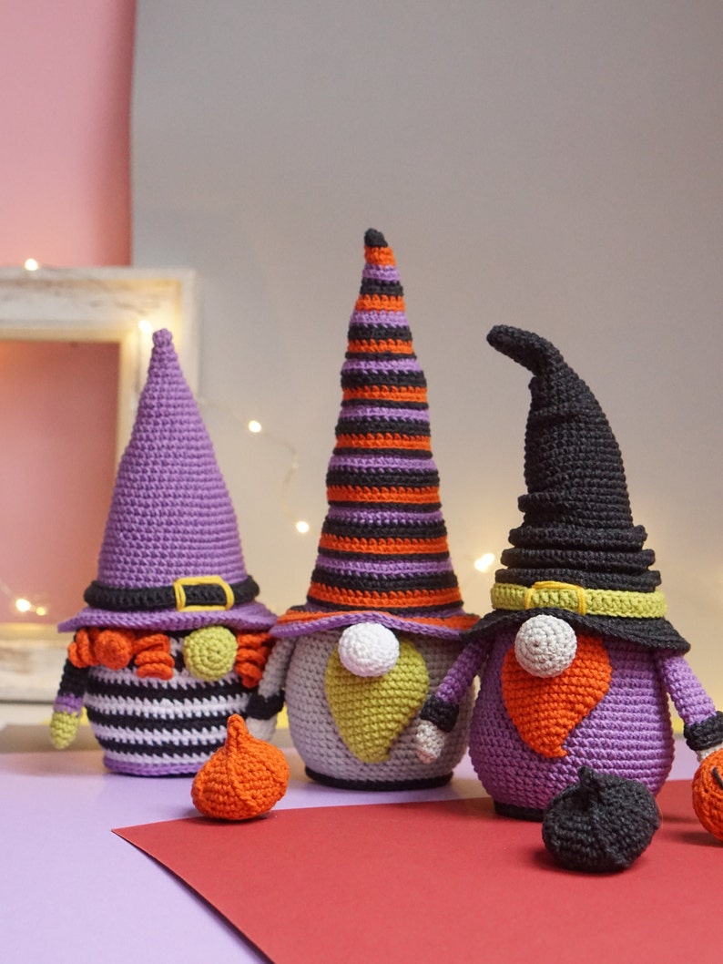 May include: Three crocheted Halloween gnomes with purple, orange, and black hats. The gnomes are holding pumpkins and are standing on a red surface.