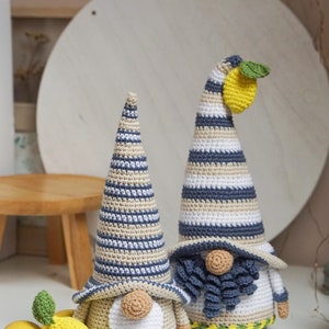 May include: Two handmade crochet gnomes with striped hats in blue, beige, and white. One gnome has a lemon and green leaf attached to its hat. A yellow lemon and a crochet lemon are next to the gnomes. The gnomes have white beards and tan noses.