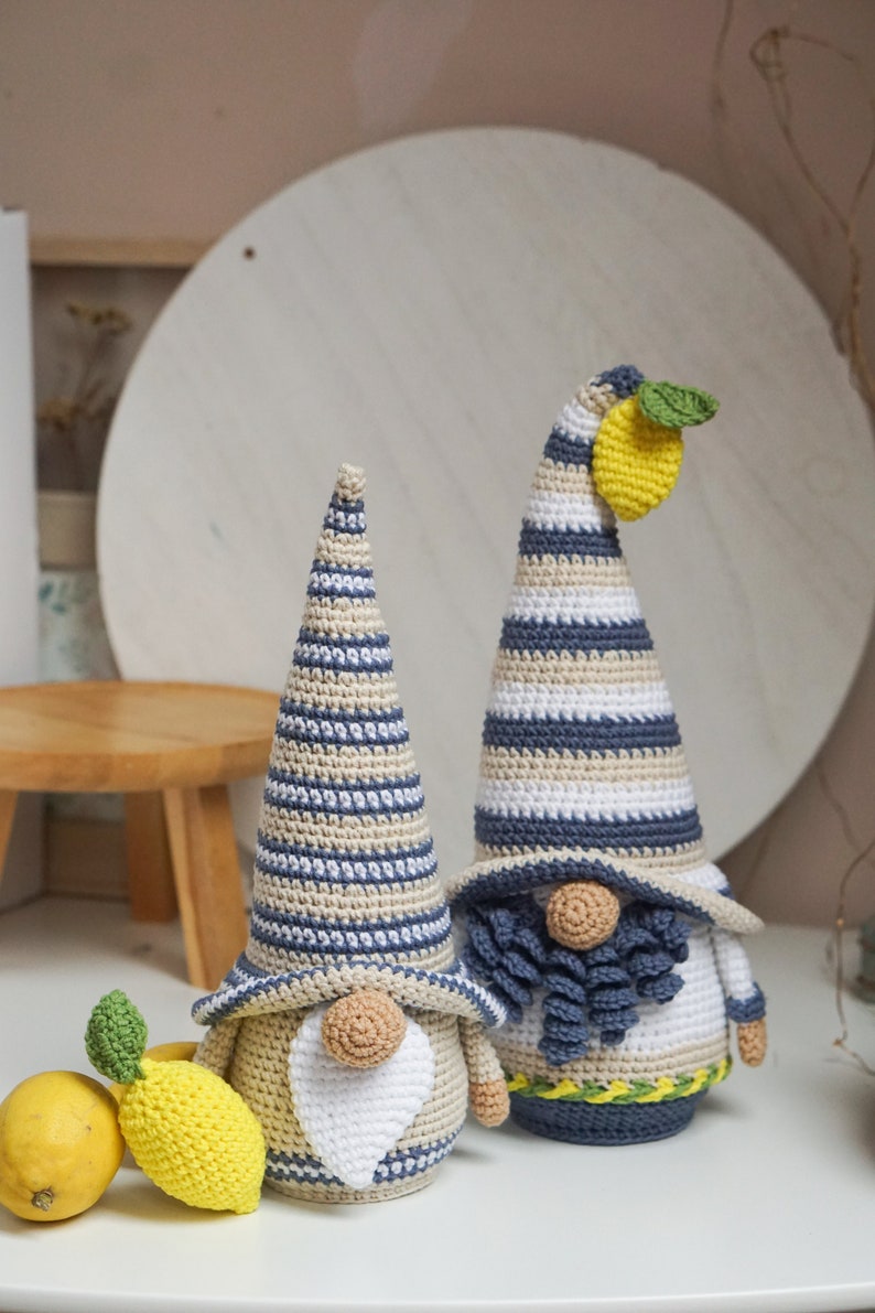 May include: Two handmade crochet gnomes with striped hats in blue, beige, and white. One gnome has a lemon and green leaf attached to its hat. A yellow lemon and a crochet lemon are next to the gnomes. The gnomes have white beards and tan noses.