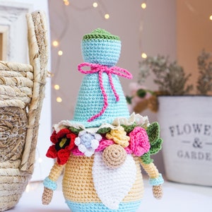 May include: A handmade crochet gnome doll with a light blue hat and body, adorned with a pink bow and a floral collar. The gnome has a white beard, and the body is yellow and blue. The doll is a decorative item.