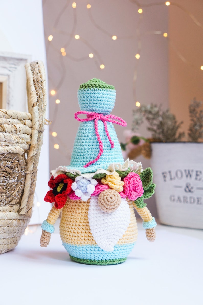 May include: A handmade crochet gnome doll with a light blue hat and body, adorned with a pink bow and a floral collar. The gnome has a white beard, and the body is yellow and blue. The doll is a decorative item.