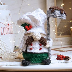 May include: A crocheted gnome wearing a white chef's hat and holding a brown and grey spatula. The gnome is wearing a green and white outfit with red accents. The gnome is sitting on a white surface.