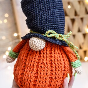 May include: A crocheted gnome with a dark blue hat, orange body, and green and yellow trim. The gnome is holding a green and white crocheted ball in its hand.