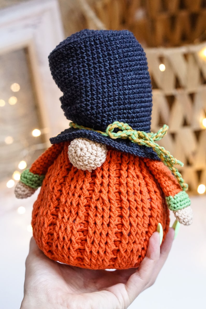 May include: A crocheted gnome with a dark blue hat, orange body, and green and yellow trim. The gnome is holding a green and white crocheted ball in its hand.
