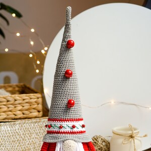 May include: A gray crocheted gnome with a red hat and beard, wearing a white and red striped band around the hat. The gnome has three red buttons on the hat and is standing on a white surface.