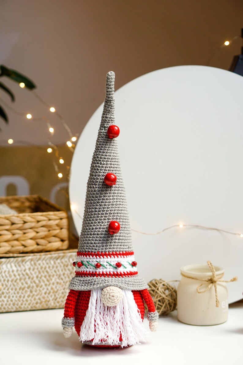 May include: A gray crocheted gnome with a red hat and beard, wearing a white and red striped band around the hat. The gnome has three red buttons on the hat and is standing on a white surface.