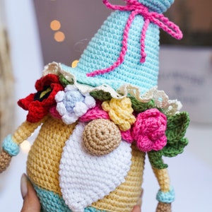 May include: A handmade crochet gnome doll with a light blue hat and a yellow body. The gnome is adorned with colorful crochet flowers, a white beard, and a pink bow. The doll is approximately 6 inches tall.