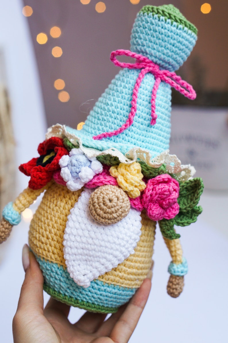 May include: A handmade crochet gnome doll with a light blue hat and a yellow body. The gnome is adorned with colorful crochet flowers, a white beard, and a pink bow. The doll is approximately 6 inches tall.