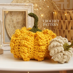 May include: Two crocheted pumpkins, one yellow and one beige, with green stems. The pumpkins are sitting on a white surface with a wooden frame and a wicker basket in the background. CROCHET PATTERN is written in the image.