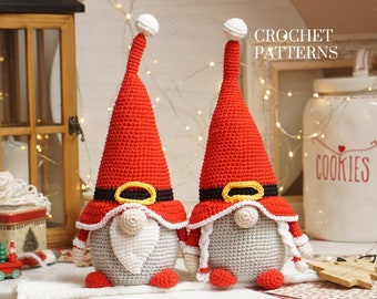 May include: Two crocheted Christmas gnomes with red hats, white beards, and yellow belts. The gnomes are sitting on a white surface with a wooden background.