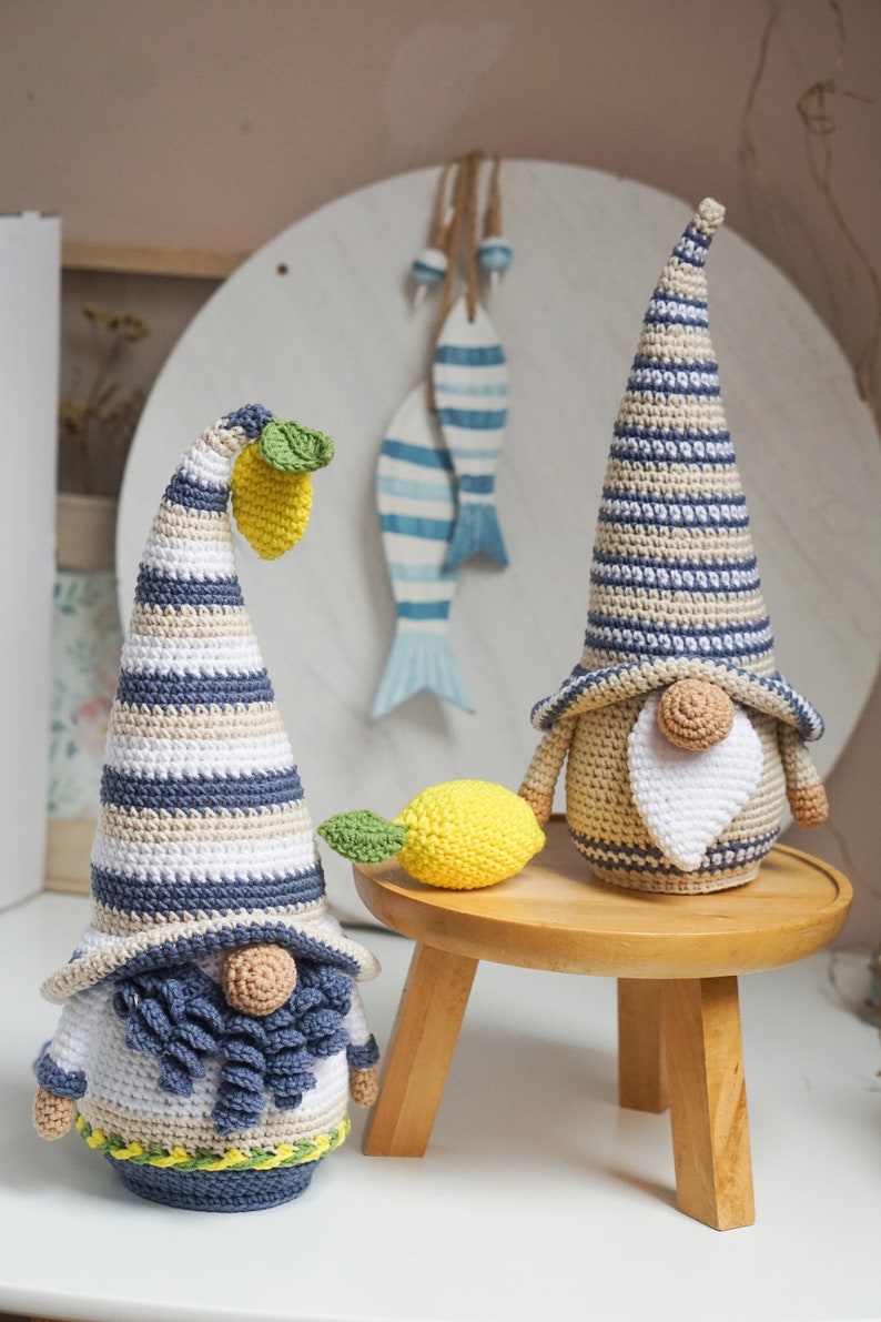 K&ouml;nnte beinhalten: Zwei geh&auml;kelte Gnome mit gestreiften H&uuml;ten und B&auml;rten. Ein Gnom hat eine gelbe Zitrone auf seinem Hut und eine gelbe Zitrone daneben. Die Gnome sitzen auf einem Holzhocker.