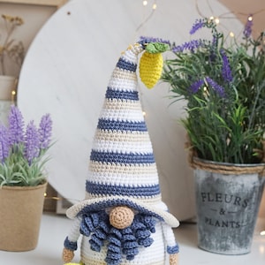 May include: Crocheted gnome figurine with a tall, striped hat in blue, white, and beige. The gnome has a beige face, blue beard, and holds a yellow lemon. A second lemon lies on the surface. The gnome is a decorative item.
