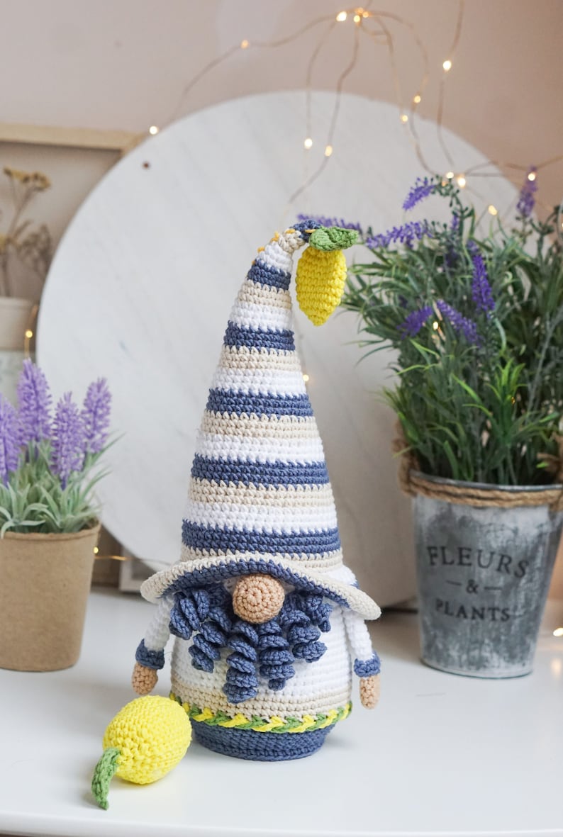 May include: Crocheted gnome figurine with a tall, striped hat in blue, white, and beige. The gnome has a beige face, blue beard, and holds a yellow lemon. A second lemon lies on the surface. The gnome is a decorative item.