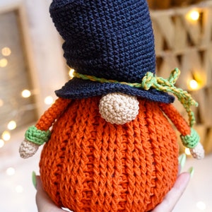 May include: A crocheted gnome with a dark blue hat, a yellow and green ribbon, and an orange body. The gnome is being held in a hand.