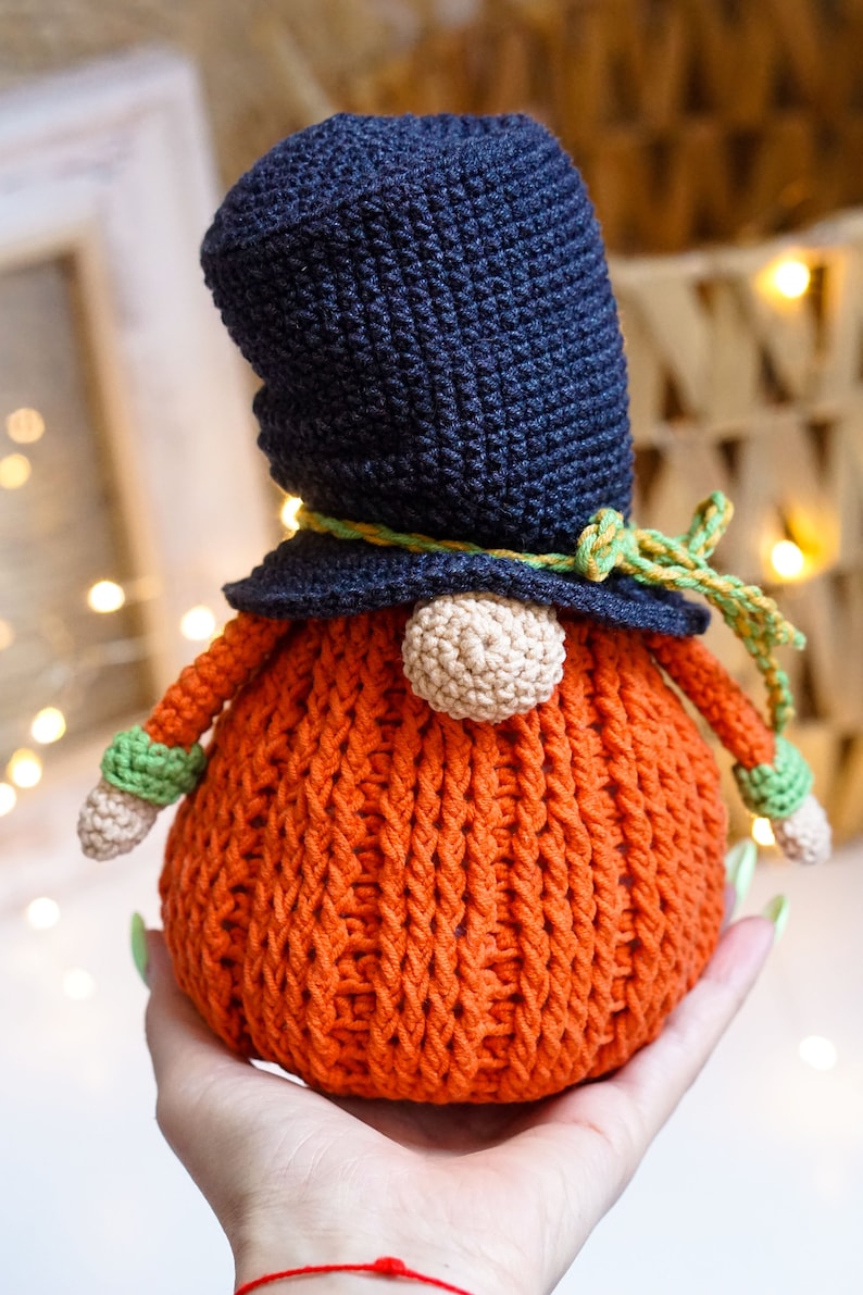 May include: A crocheted gnome with a dark blue hat, a yellow and green ribbon, and an orange body. The gnome is being held in a hand.
