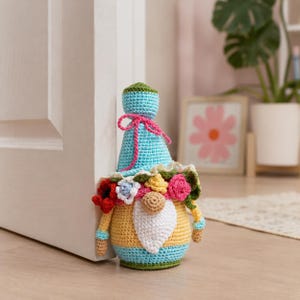 Cute flower gnome used as a functional crochet door stopper. Weighted amigurumi plush gnome holding the door open, unique and practical handmade home accessory