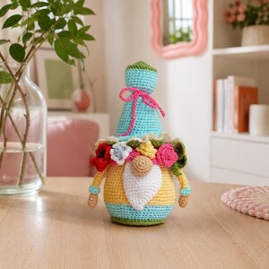 May include: A handmade crochet gnome doll with a blue hat tied with a pink bow. The gnome has a yellow body, a white beard, and is adorned with colorful flowers. The doll is sitting on a wooden table.